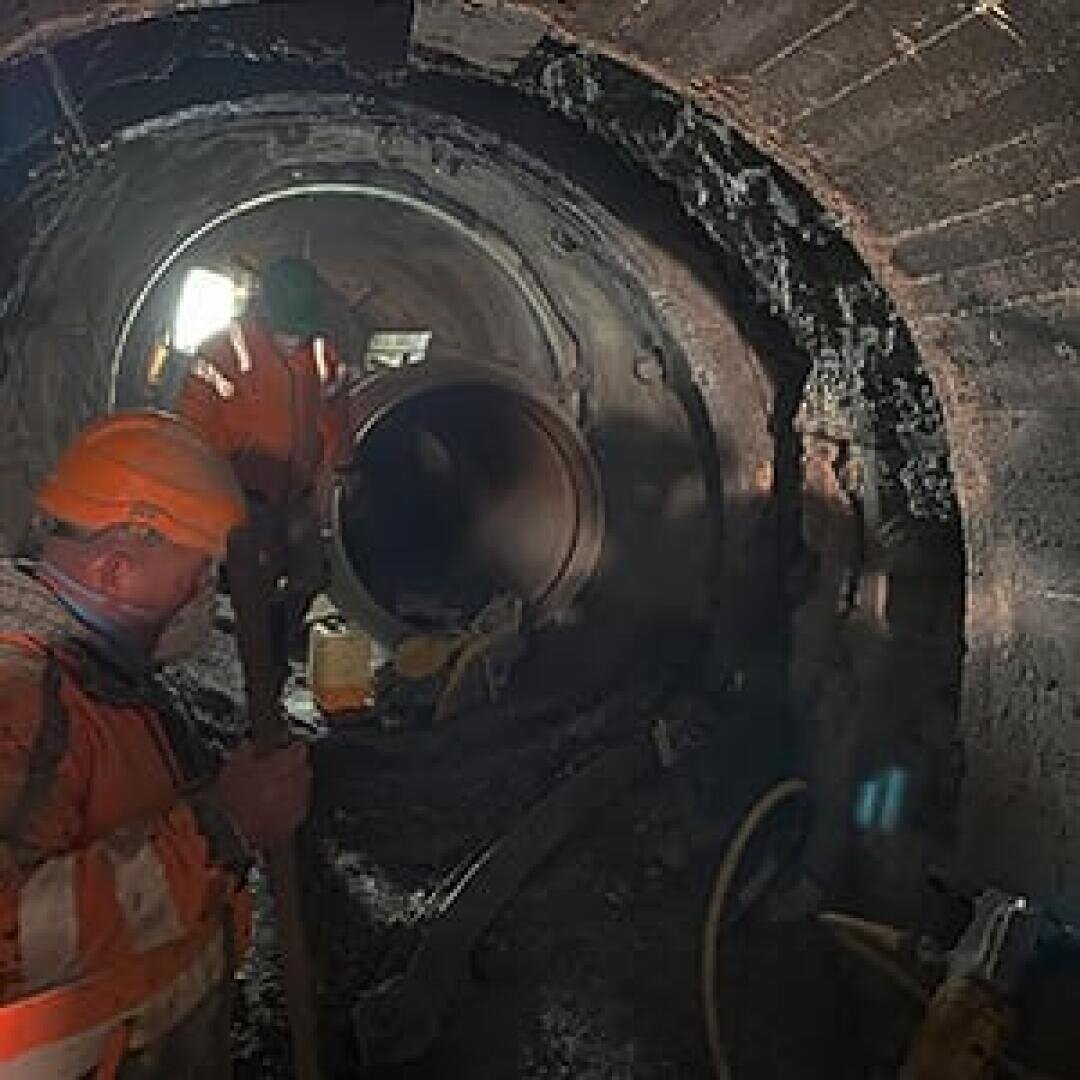 Zwei Bauarbeiter, die orangefarbene Schutzkleidung und Helme tragen, arbeiten in einem schwach beleuchteten, großen unterirdischen Rohrtunnel. Einer hält ein Werkzeug in der Hand, der andere steht in der Nähe eines großen Metallrohrs. Licht scheint vom Tunneleingang herein.