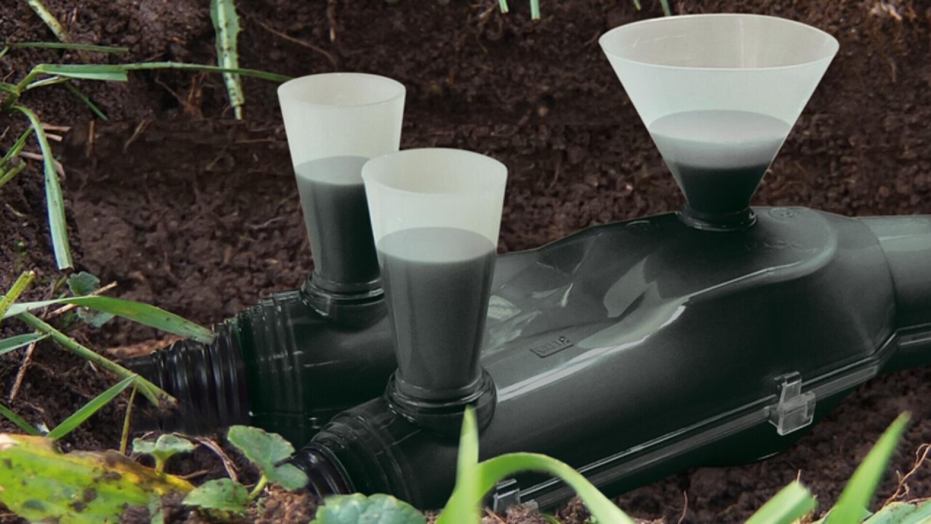 A black pipe partially buried in soil with three plastic funnels inserted on top, filled with dark liquid. Green grass and plants surround the pipe in an outdoor setting.