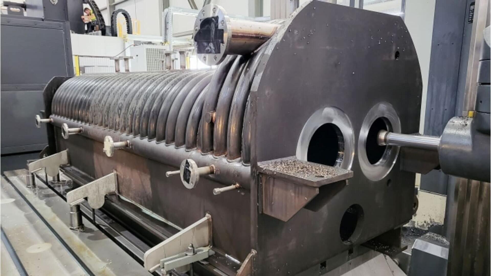 Large industrial cylindrical metal machine with curved pipes along the top, two circular openings at the front, and mounted on a factory floor with gray equipment and wires visible in the background.