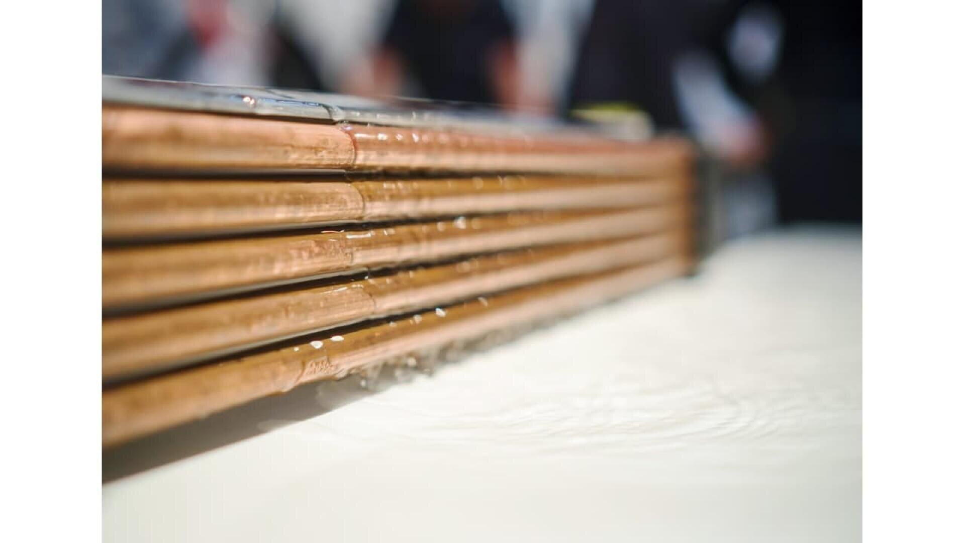 Close-up of several copper pipes with water trickling down their sides, placed over a smooth, white surface. The background is blurred, with indistinct dark shapes.