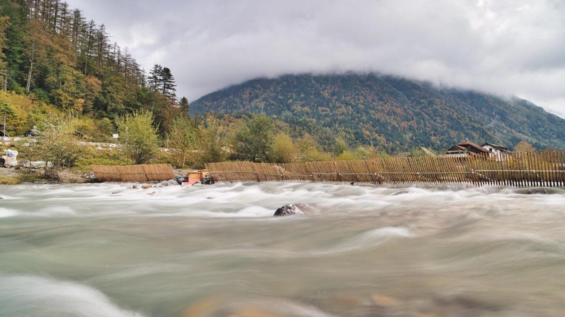 A fast-flowing river bordered by wooden fences runs through a lush valley with green trees. In the background, a forested mountain rises under a cloudy sky. A few buildings are nestled near the riverbank.