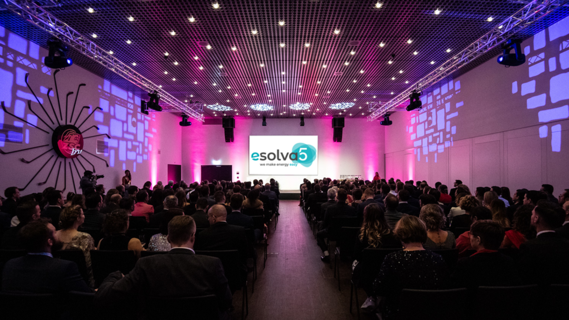 Ein großes Publikum in förmlicher Kleidung sitzt in einem modernen, violett beleuchteten Veranstaltungssaal vor einer Bühne mit einer Leinwand, auf der das "esolva"-Logo zu sehen ist. Dekorative Lichter und Wandgrafiken schaffen eine elegante Atmosphäre.