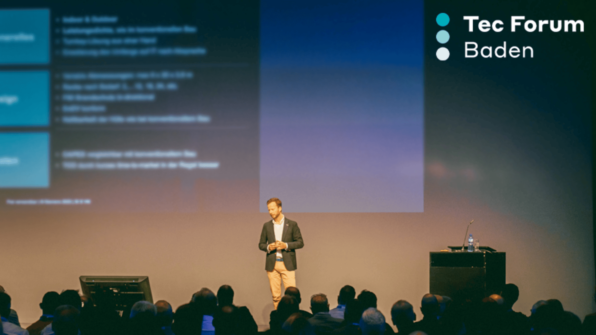 Ein Mann im Blazer steht auf der Bühne und hält einen Vortrag vor dem Publikum des Tec Forum Baden; hinter ihm werden Dias projiziert, und in der rechten oberen Ecke ist das Logo der Veranstaltung zu sehen.