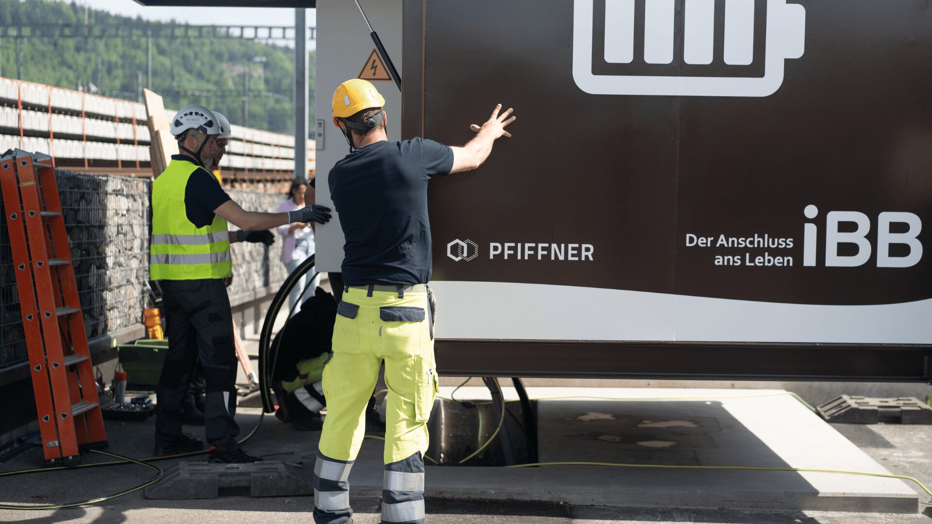 Zwei Bauarbeiter in Schutzkleidung stellen im Freien Geräte in der Nähe eines großen braunen Containers mit der Aufschrift PfiFFNER und iBB auf. Ein Arbeiter zeigt auf etwas, während der andere etwas neben einer orangefarbenen Leiter justiert.