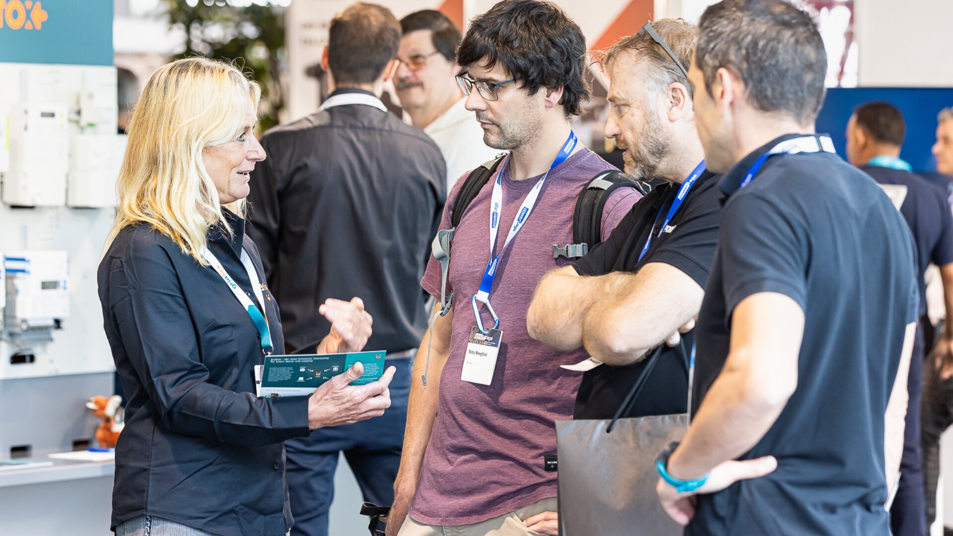 Eine Frau spricht auf einer Konferenz oder Messe mit drei Männern. Sie stehen zusammen und unterhalten sich, alle tragen Namensschilder und Freizeitkleidung. Stände und andere Teilnehmer sind im unscharfen Hintergrund zu sehen.