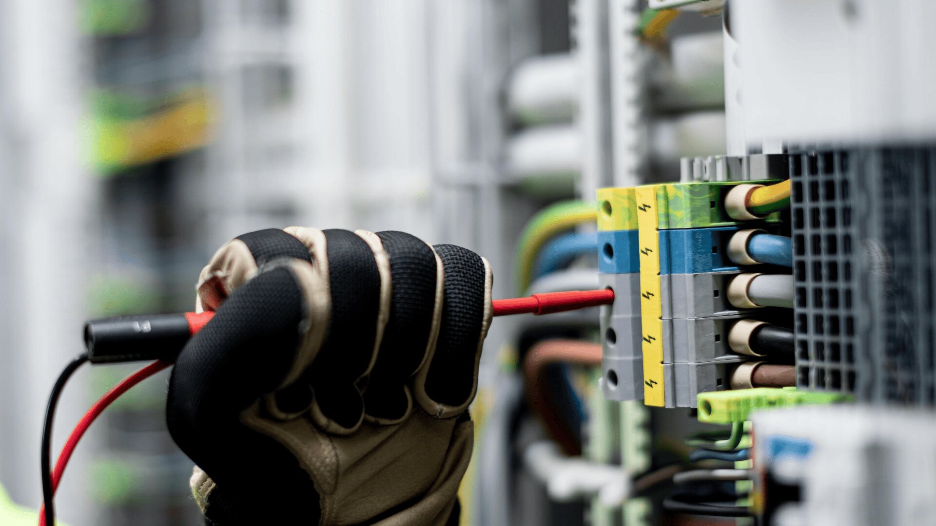 Eine behandschuhte Hand hält eine rote Sonde, mit der sie die Verdrahtung und die Komponenten in einer elektrischen Schalttafel mit farbigen Kabeln und Sicherungen prüft oder inspiziert.