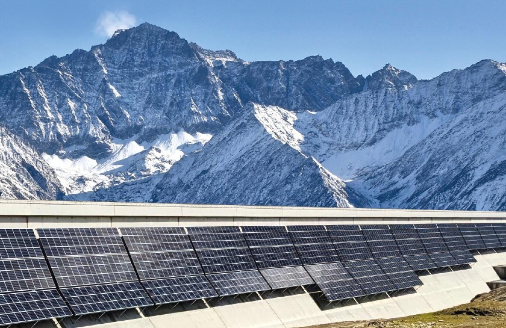 Rows of solar panels on a concrete platform are set against a backdrop of high, snow-capped mountains under a bright blue sky.