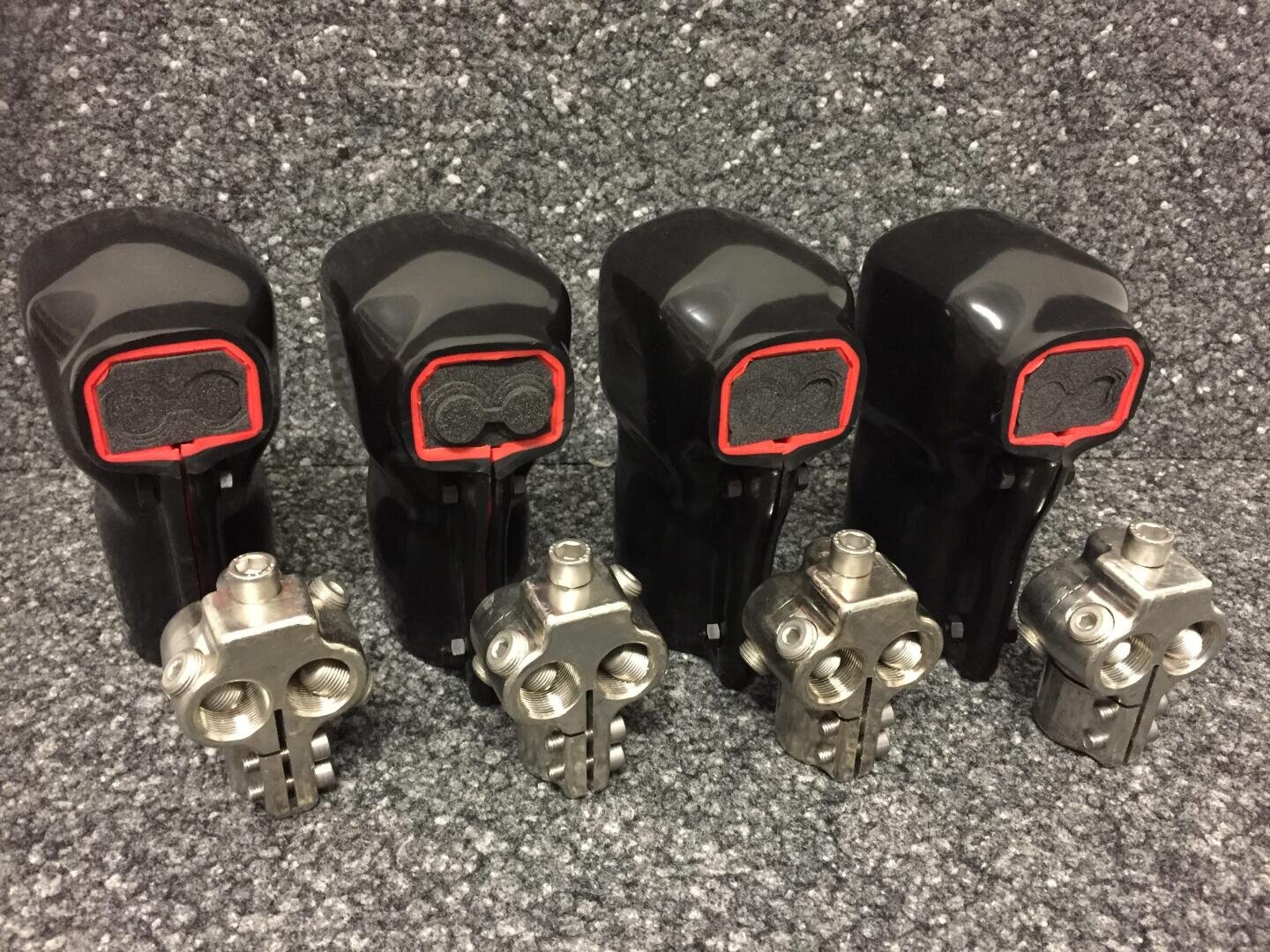 Vier schwarze Joystick-Bedienelemente mit roten Umrissen sind in einer Reihe hinter vier mechanischen Metallkomponenten angeordnet, die alle auf einer grauen, strukturierten Oberfläche liegen.