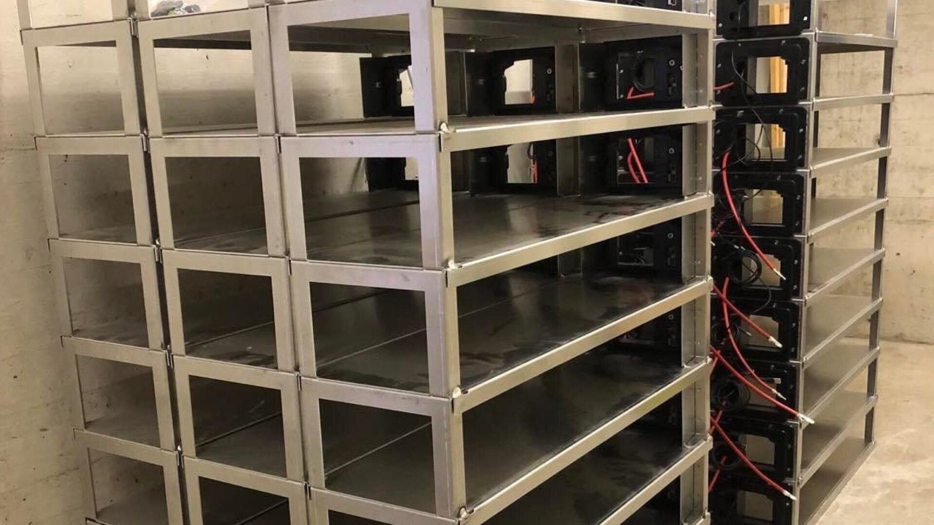 Metallregale mit leeren Fächern sind vor einer Betonwand gestapelt. Einige Einheiten haben freiliegende rote und schwarze Kabel, was auf eine mögliche Verwendung für Elektronik oder Datenserver hindeutet. Der Raum wirkt industriell und unfertig.