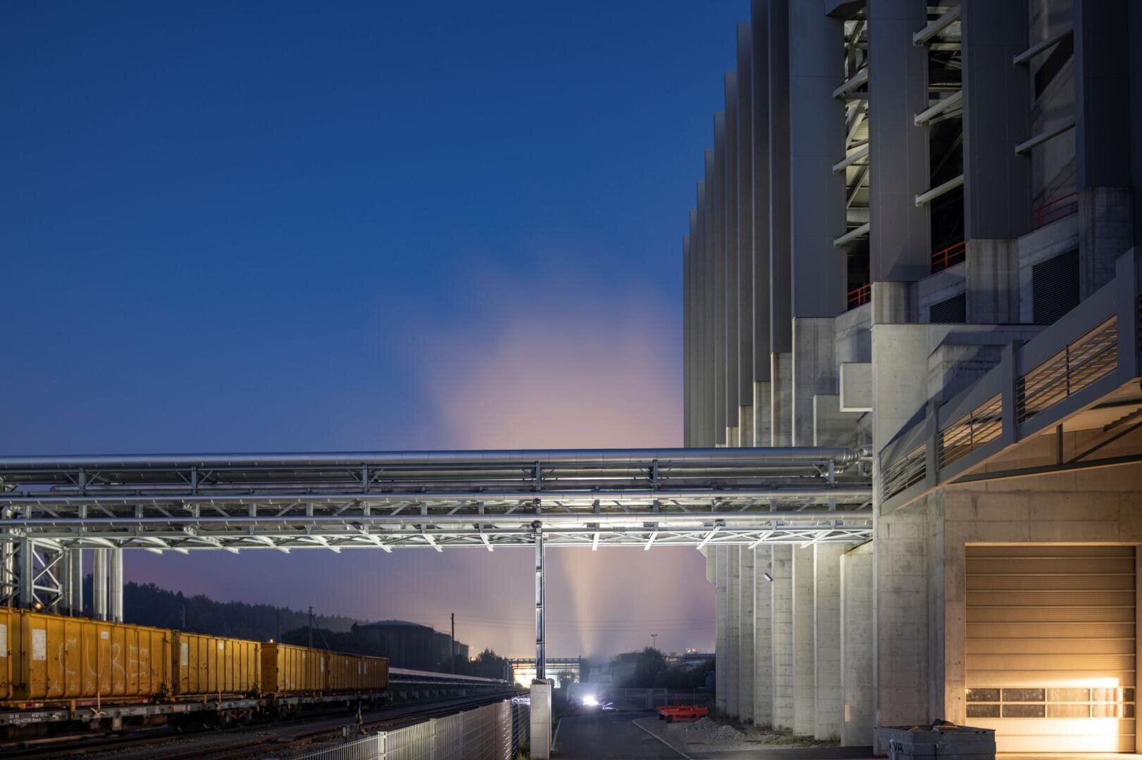A modern industrial plant at dusk with large concrete structures, metal pipes and yellow freight train wagons on the tracks. Bright lights illuminate the scene and fog or smoke rises in the background.