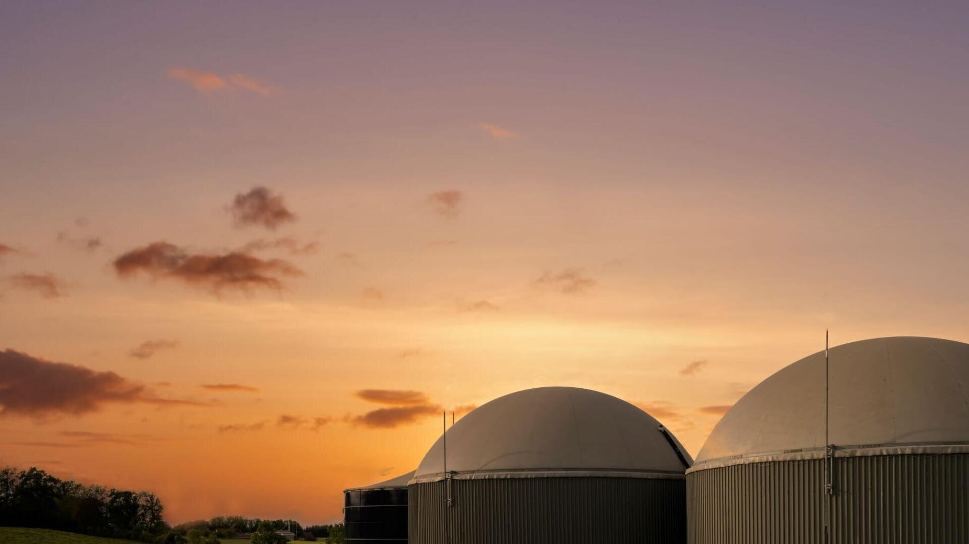 Zwei große, kuppelförmige Biogasanlagen vor einem farbenfrohen Sonnenuntergangshimmel mit vereinzelten Wolken und einer Andeutung von Bäumen am Horizont.