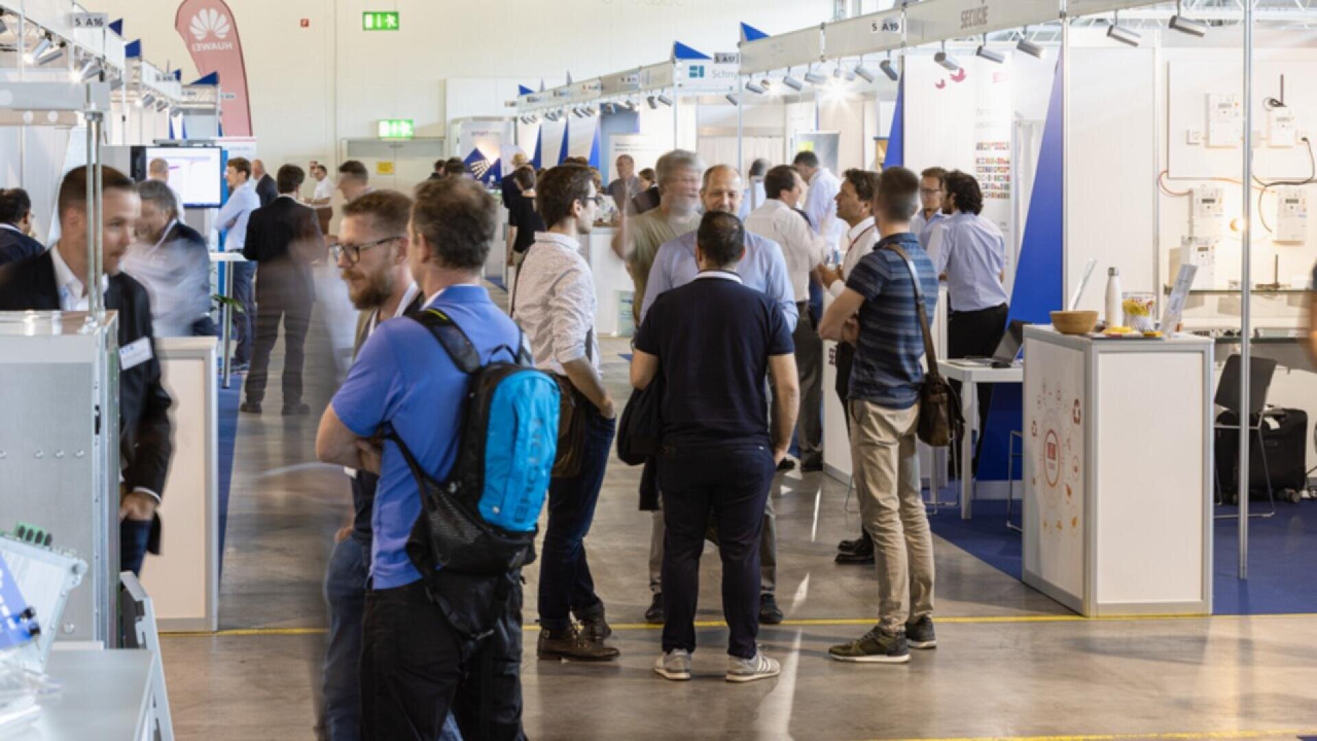People are gathered and conversing in a large exhibition hall with booths and displays along both sides. Some attendees hold brochures and wear backpacks, creating a busy, professional atmosphere.