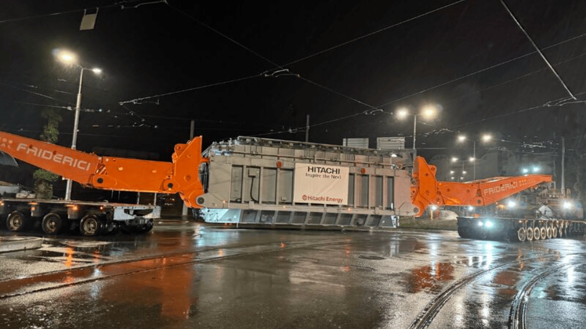 Ein großer Industrietransformator mit der Aufschrift Hitachi Energy wird nachts auf einem Spezialanhänger mit orangefarbenen Stützen transportiert. Die Straße ist nass und reflektiert in der Dunkelheit die Straßenlaternen.
