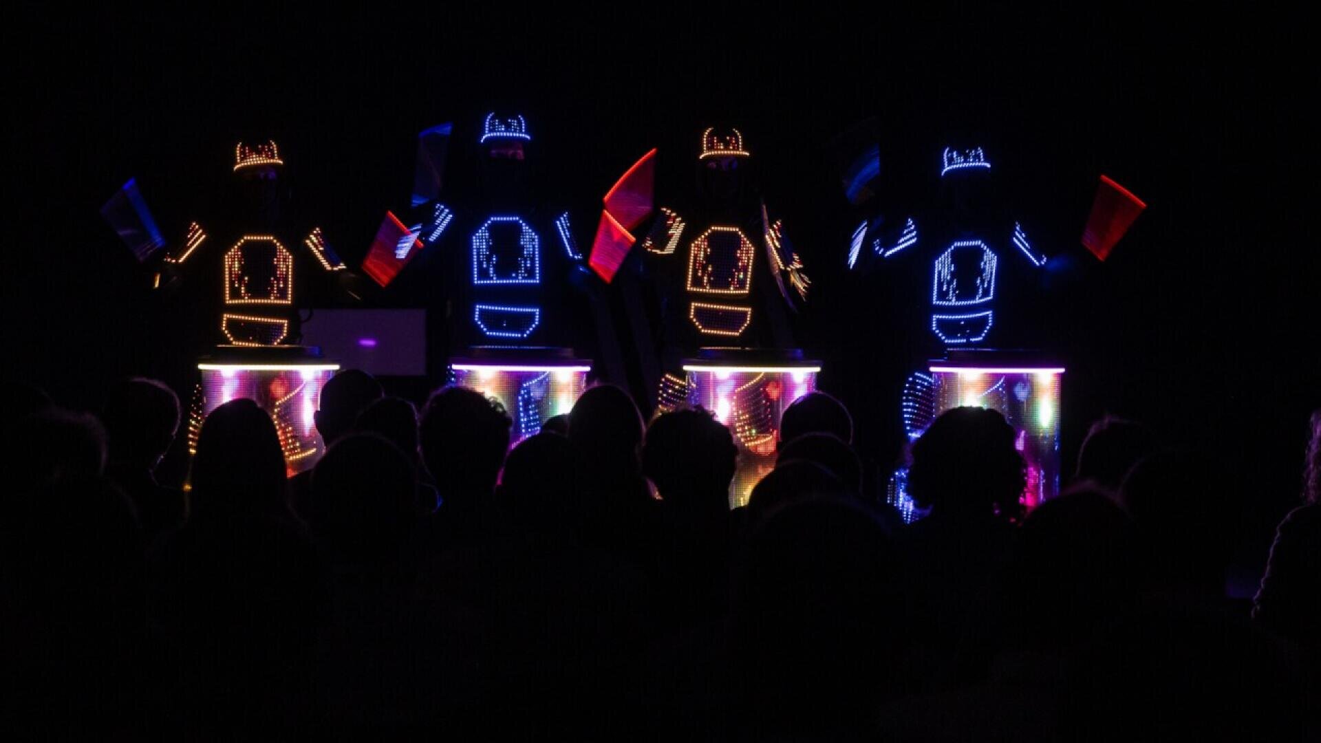 Five performers in dark suits with glowing neon lights play illuminated drums on a dark stage, creating a vibrant, colorful light show for a silhouetted audience.