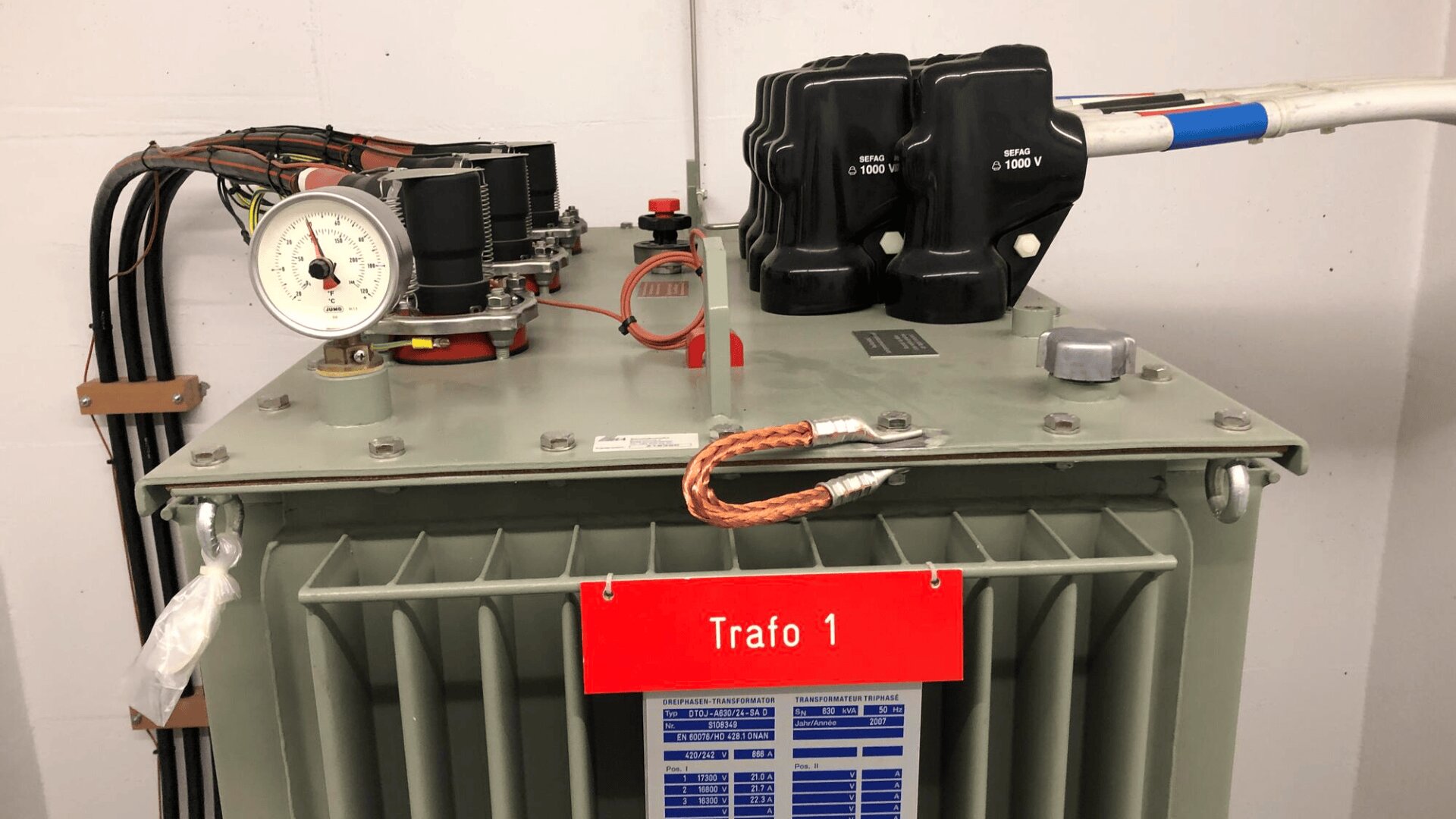 Ein grauer elektrischer Transformator mit der Bezeichnung Trafo 1 mit angeschlossenen Kabeln, Schaltern, einem Messgerät und einem roten Schild. An der Vorderseite des Transformators befinden sich technische Informationsschilder.