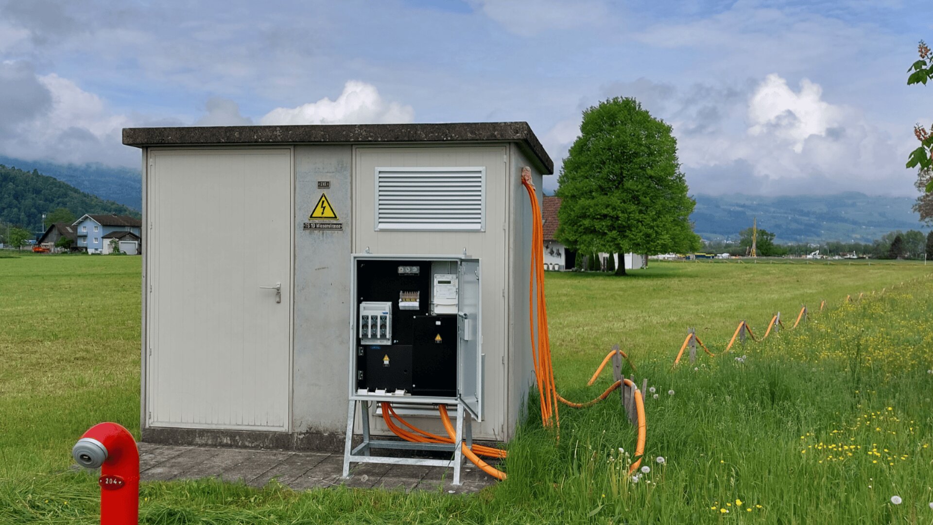 Ein Stromkasten mit offenen Schalttafeln und orangefarbenen Kabeln steht in einem grasbewachsenen Feld, mit Hügeln und bewölktem Himmel im Hintergrund. Im Vordergrund ist ein roter Hydrant zu sehen und in der Nähe wachsen gelbe Wildblumen.
