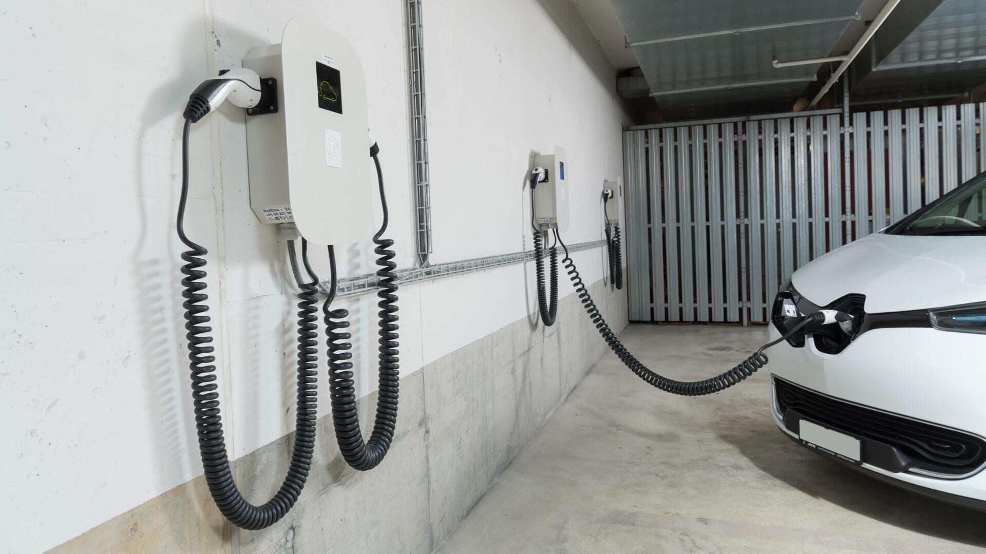 Ein weißes Elektroauto ist in einer Tiefgarage geparkt und mit einem Spiralkabel an eine an der Wand befestigte Ladestation angeschlossen. Weitere Ladestationen sind entlang der Wand zu sehen.