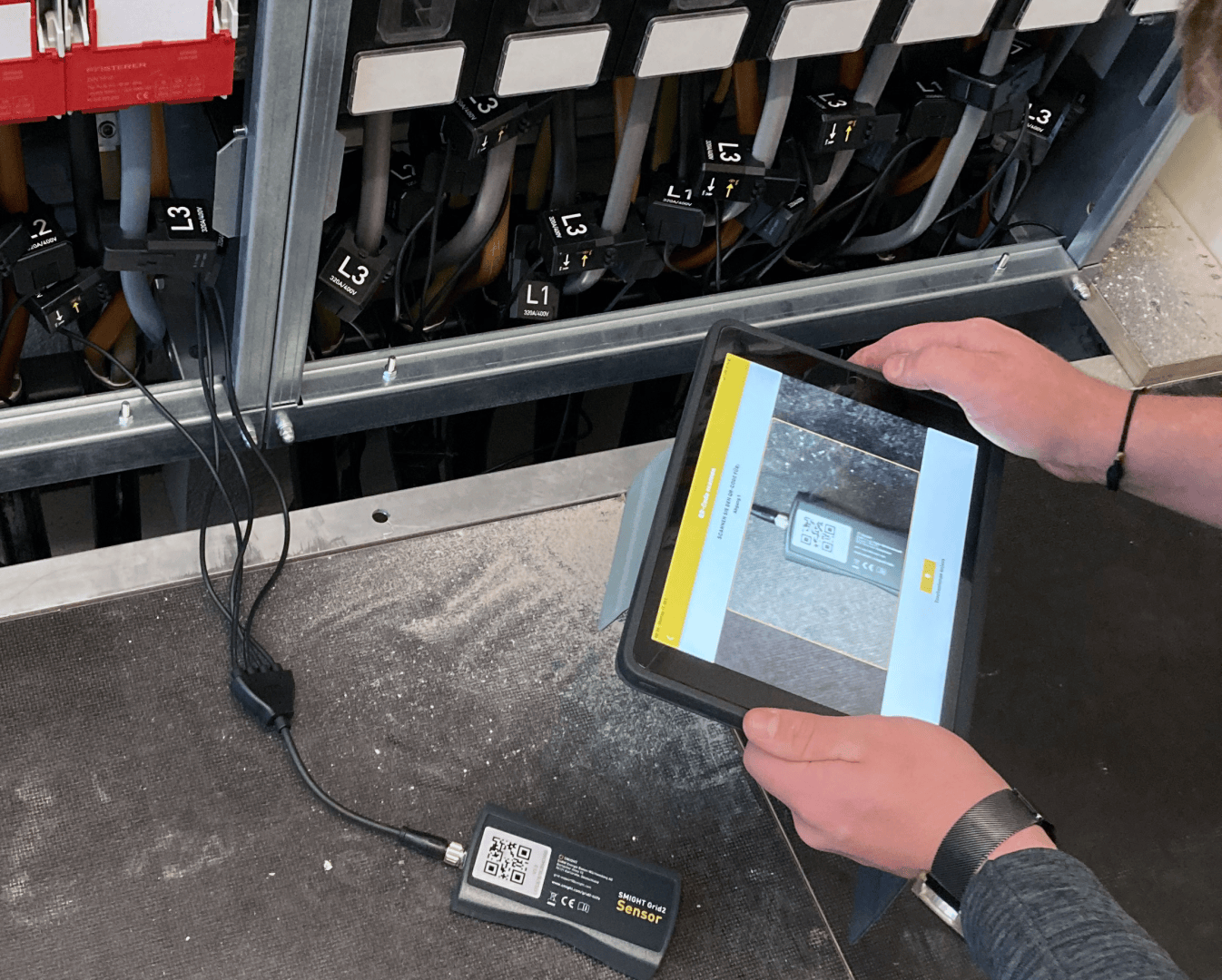 Eine Person hält ein Tablet mit einem QR-Code, das mit einem kompakten Gerät verbunden ist, vor eine elektrische Anlage mit beschrifteten Kabeln und Schaltern.