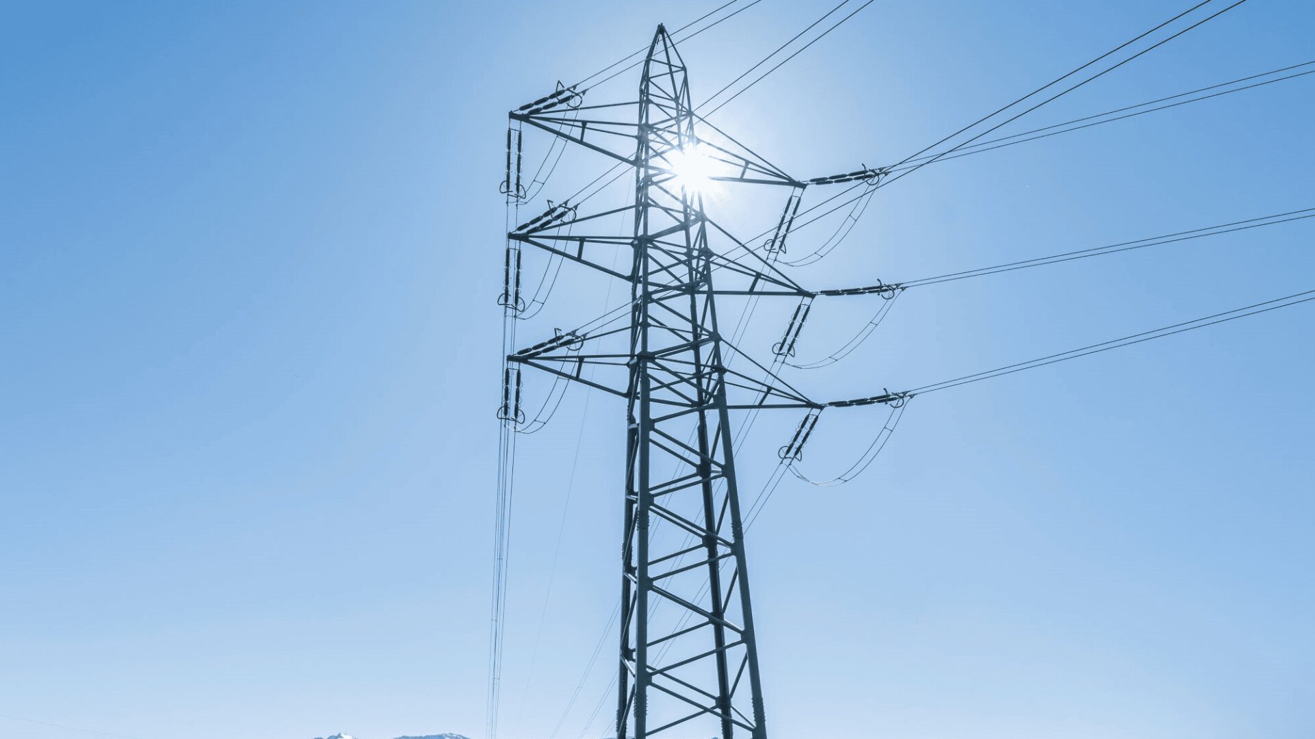 Ein hoher metallener Strommast mit Stromleitungen ragt in den blauen Himmel, und hinter dem Bauwerk scheint die helle Sonne.