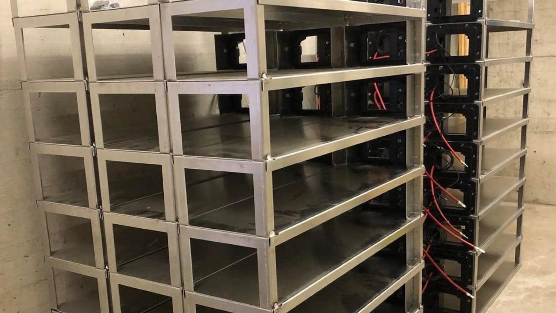A stack of empty metal shelves or racks is positioned against a concrete wall in an industrial-looking room. Red and black wires are visible on the right side of the racks.