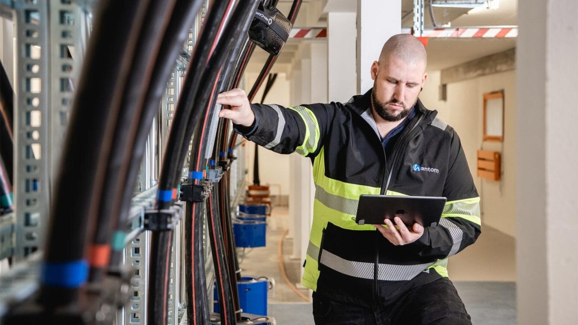 A man in a high-visibility jacket stands in an industrial setting, holding onto a cable rack with one hand and using a tablet with the other, appearing focused on his work.