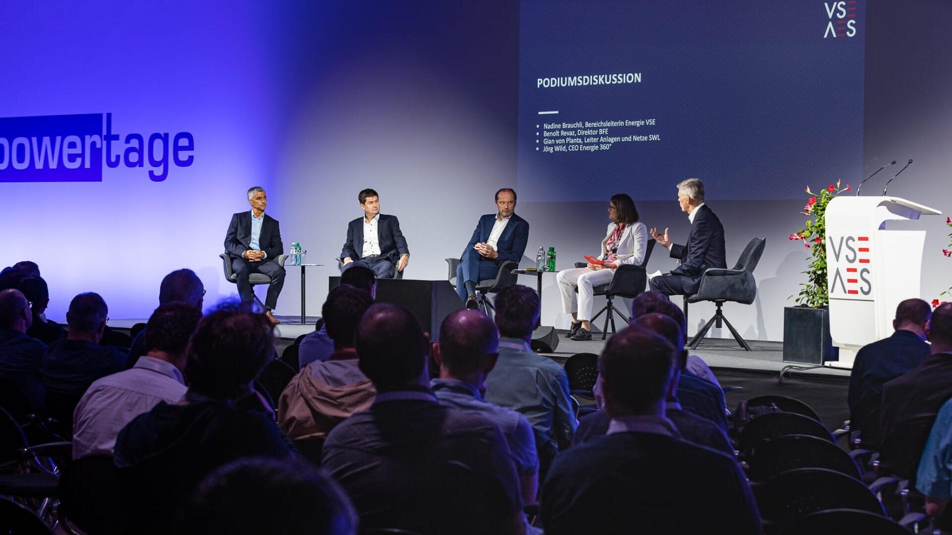 Eine Gruppe von fünf Personen sitzt auf einer Konferenz auf einer Bühne und diskutiert mit Blick auf das Publikum. Auf einer Präsentationsfolie hinter ihnen sind Namen und Titel aufgeführt. Die Bühne ist gut beleuchtet, und das Branding der Veranstaltung ist an den Wänden sichtbar.