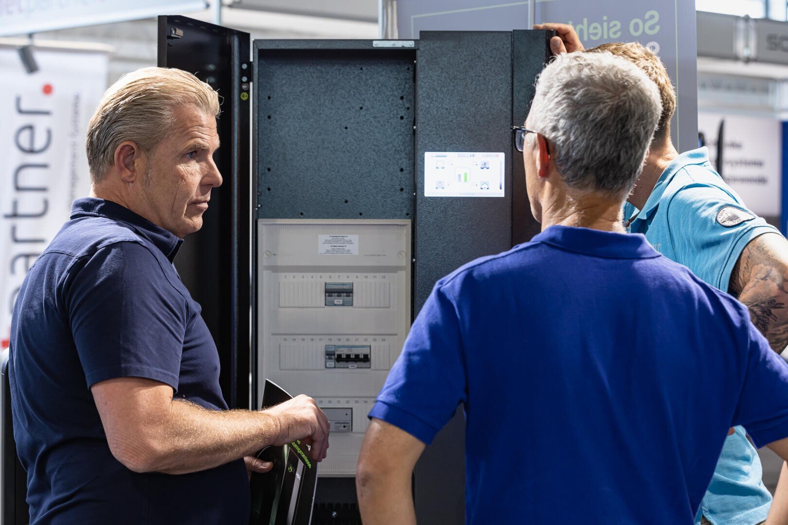 Drei Männer stehen vor einer offenen elektrischen Schalttafel und diskutieren über deren Bestandteile. Ein Mann zeigt auf die Schalttafel, während die anderen aufmerksam zuhören. Sie scheinen auf einer professionellen Veranstaltung oder Messe zu sein.
