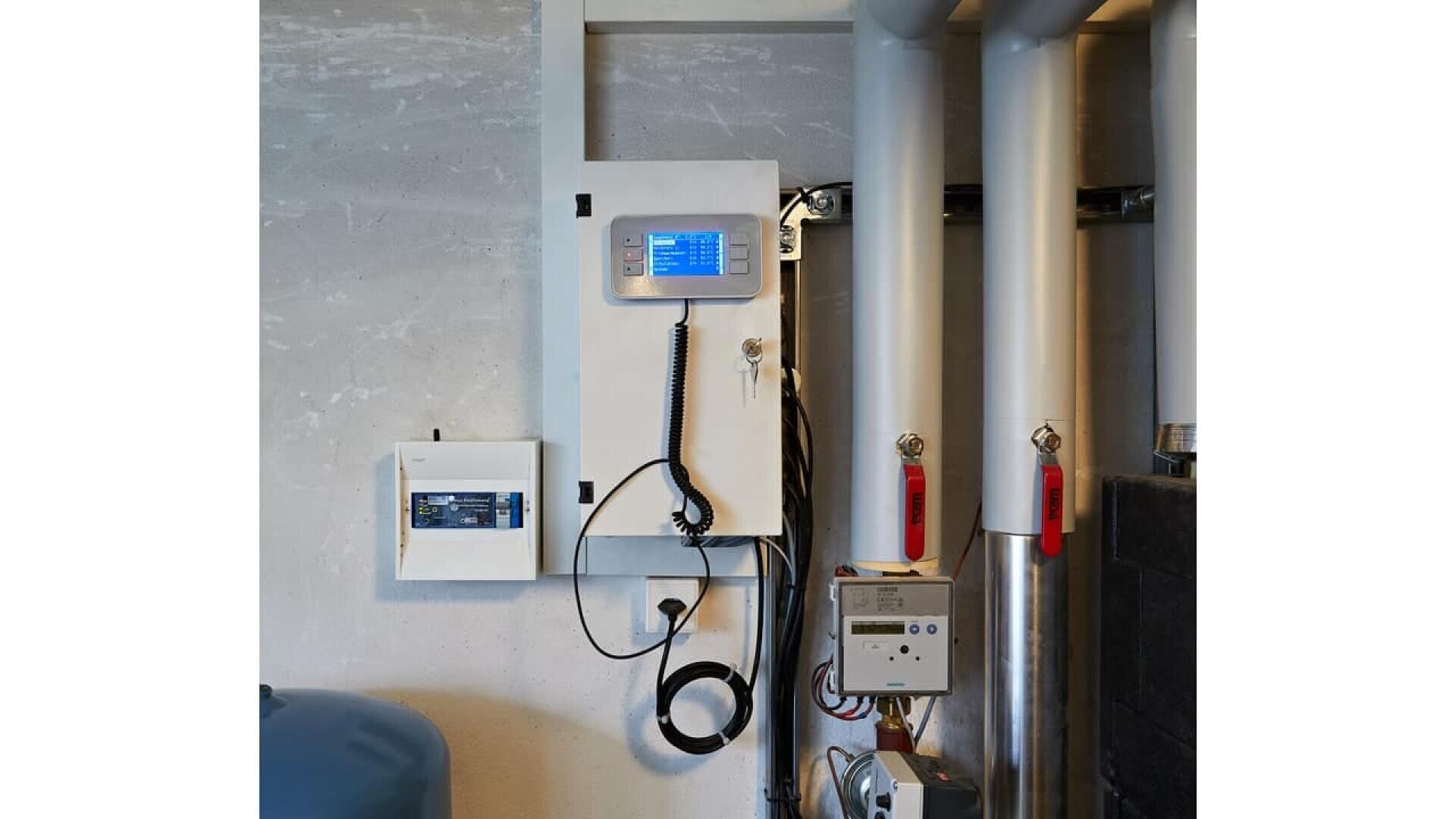 A control panel with a screen and coiled cord is mounted on a wall next to pipes with red valves and other control boxes in an industrial or utility room.