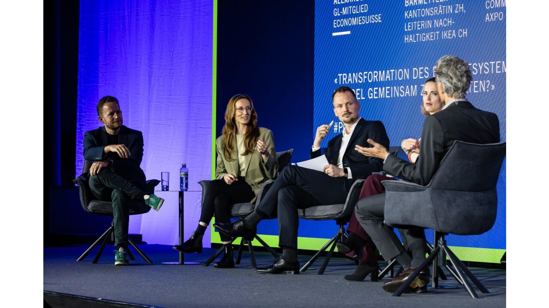 Five people in business attire sit on stage in chairs, engaged in a panel discussion. A blue backdrop with text and a green floor border are visible behind them. One person gestures while others listen attentively.