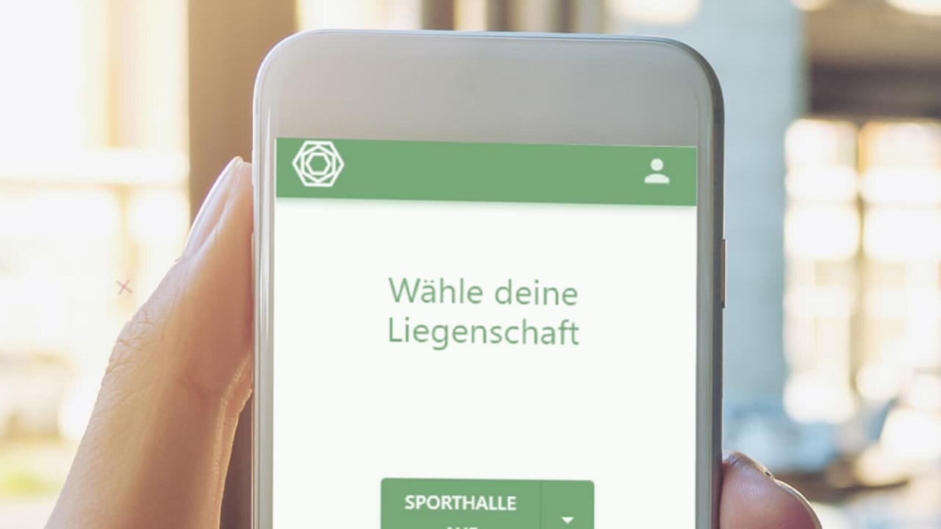 A hand holding a smartphone displaying a German app with the text Wähle deine Liegenschaft and a green button labeled Sporthalle on the screen. The background is softly blurred.