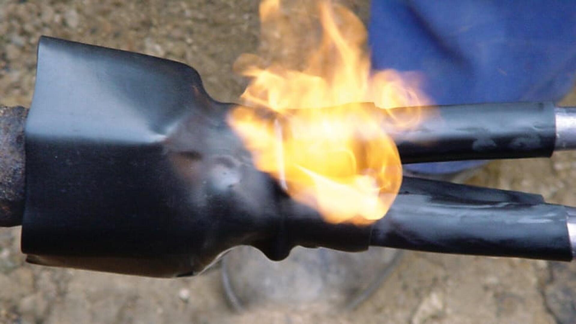 Ein Schrumpfschlauch wird mit einer offenen Flamme auf ein elektrisches Kabel aufgebracht, wodurch der Schlauch fest um die Verbindung schrumpft. Der Hintergrund ist unscharf.