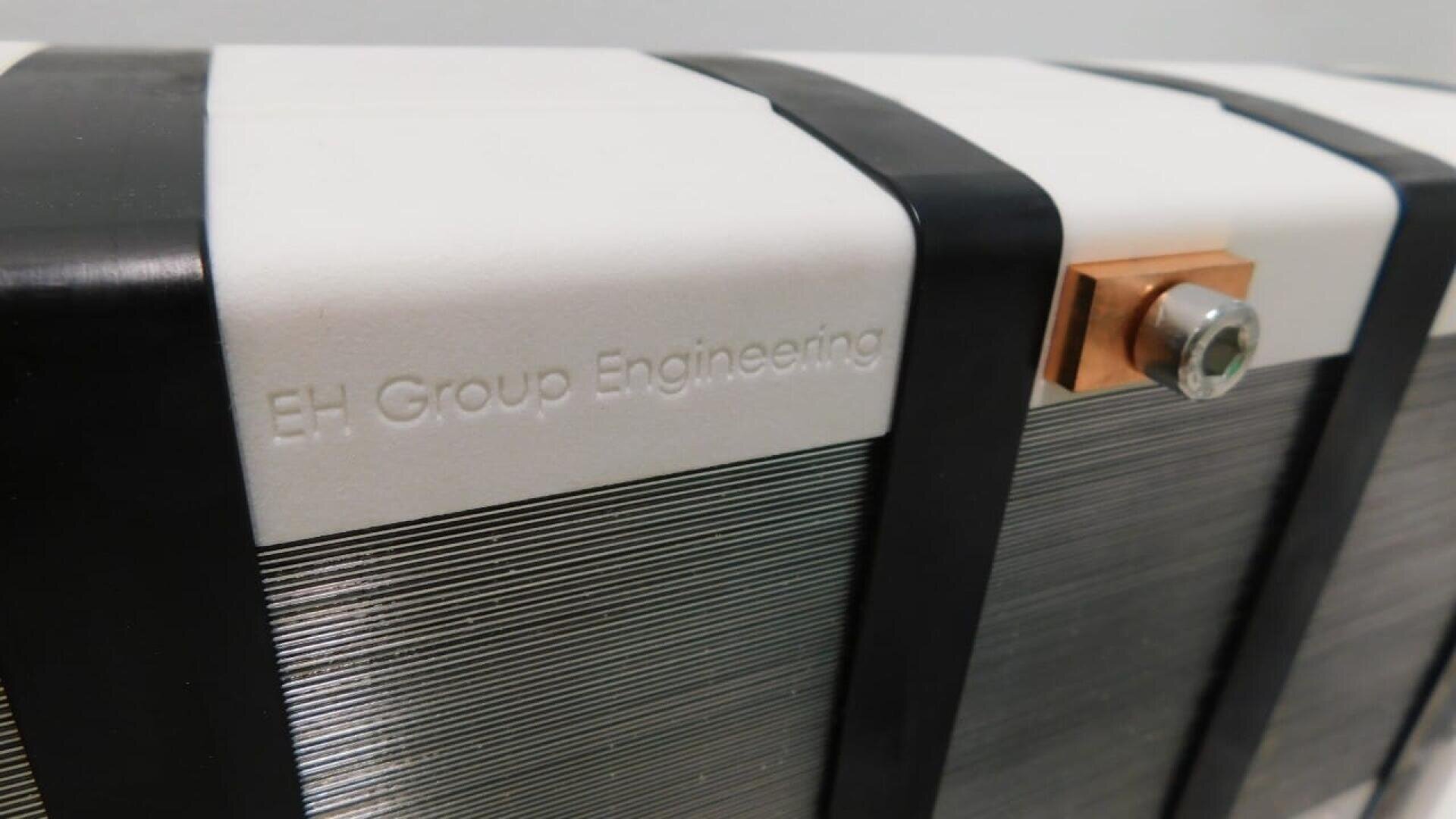 Nahaufnahme eines Geräts mit der Prägung EH Group Engineering auf einer weißen Fläche, mit Metallplatten, schwarzen Stützbändern und einem Kupferverbinder, der mit einem Bolzen befestigt ist.