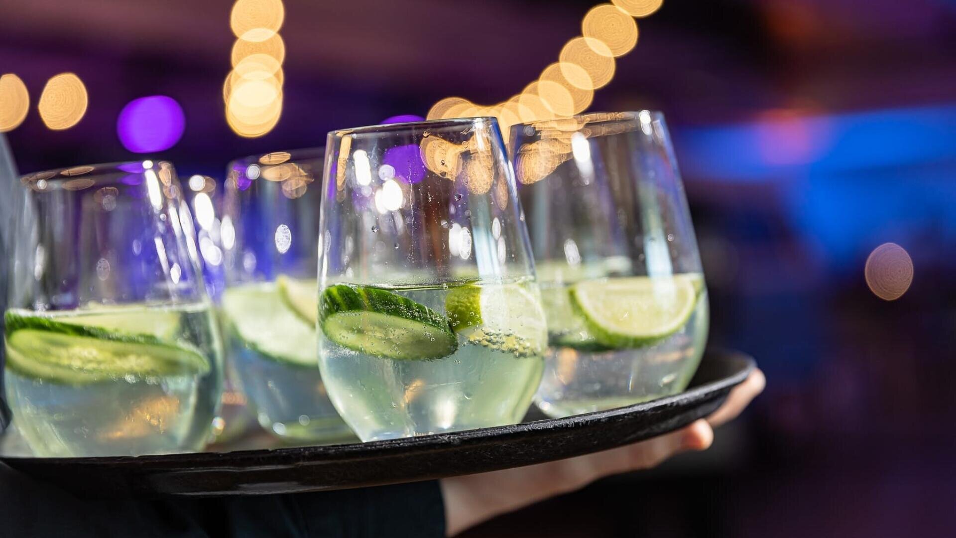 Vier Gläser mit klaren Cocktails, die mit Limetten- und Gurkenscheiben garniert sind, werden auf einem schwarzen Tablett serviert, wobei verschwommene Lichterketten und ein violett-blauer Hintergrund einen festlichen Rahmen suggerieren.