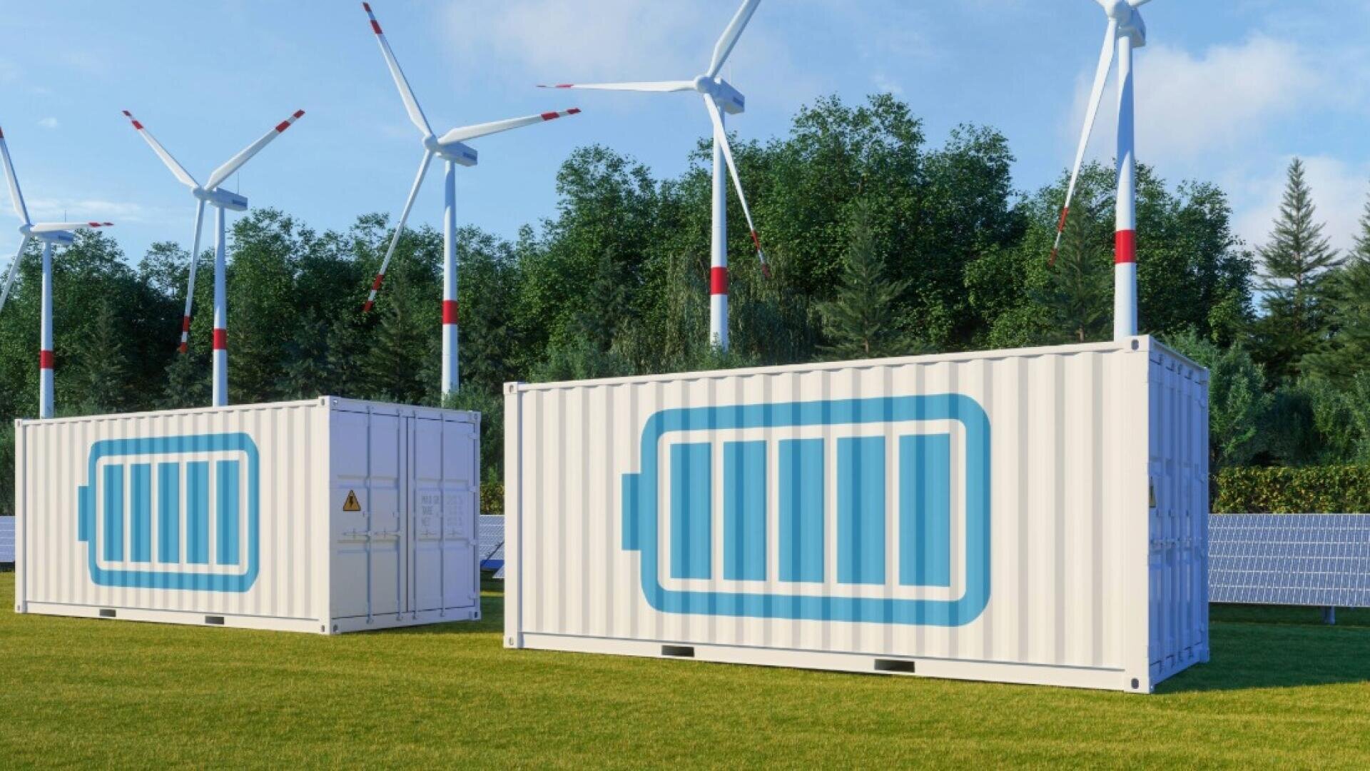 White shipping containers with large battery symbols stand on grass, with wind turbines and solar panels in the background, near a line of green trees under a blue sky.