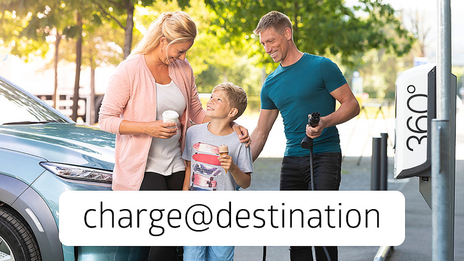 Eine dreiköpfige Familie lächelt gemeinsam in der Nähe einer Ladestation für Elektrofahrzeuge im Freien. Die Mutter und der Sohn halten Getränke, der Vater einen Ladestecker. Der Text charge@destination ist über dem Bild eingeblendet.