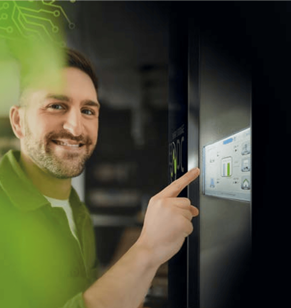 Ein lächelnder Mann berührt ein digitales Bedienfeld an einer dunklen Maschine mit grünen, schaltkreisähnlichen Grafiken und Pflanzenblättern, die leicht im Vordergrund zu erkennen sind.