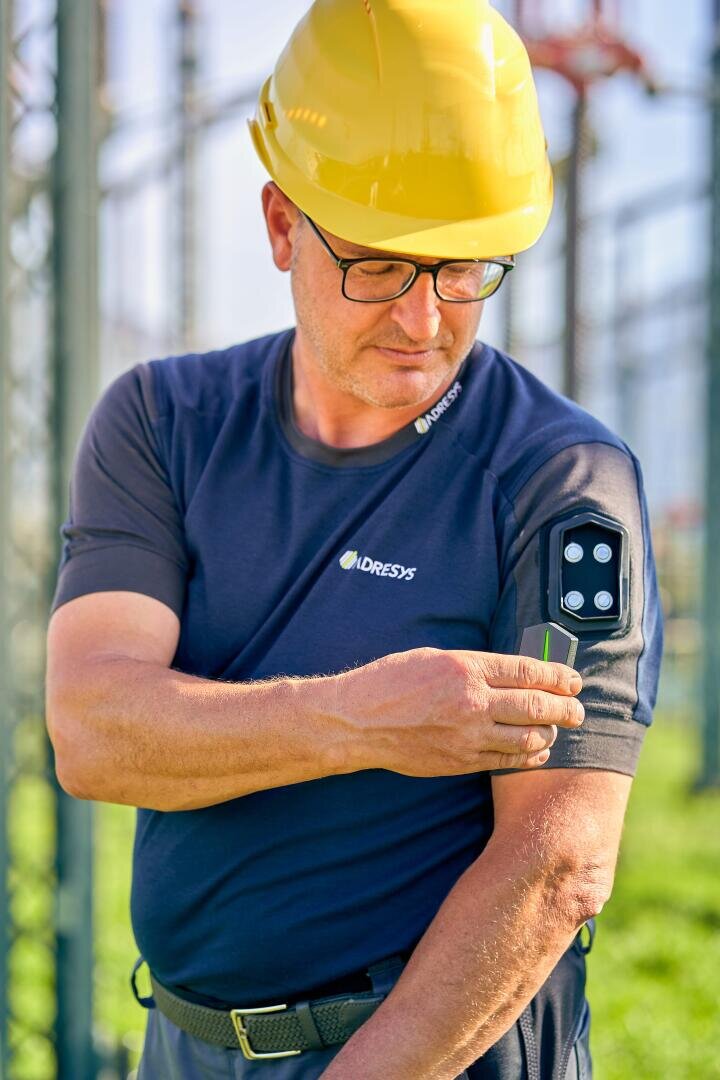 Ein Mann mit einem gelben Schutzhelm und einer Brille steht im Freien und schaut auf ein schwarzes Gerät, das am Ärmel seines Hemdes befestigt ist. Er berührt das Gerät mit einer Hand.