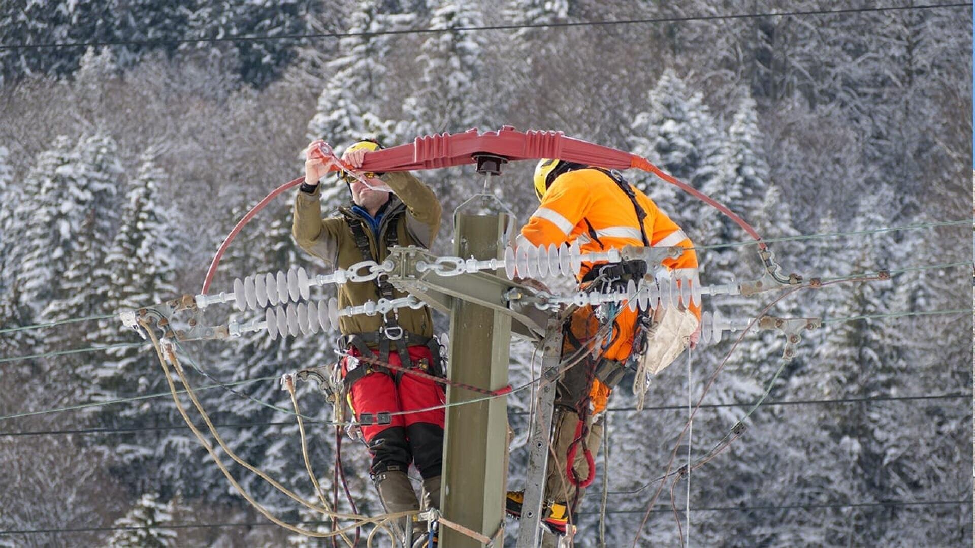 Zwei Arbeiter eines Versorgungsunternehmens in Schutzkleidung reparieren Stromleitungen auf einem hohen Strommast, umgeben von schneebedeckten Bäumen und einer winterlichen Landschaft im Hintergrund.