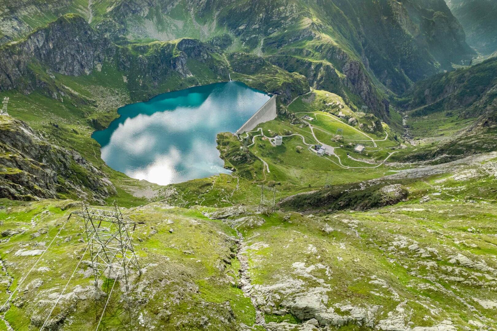 Aerial view of a turquoise mountain lake surrounded by green hills and rocky terrain, with a dam, winding roads, power lines, and scattered buildings under a partly cloudy sky.
