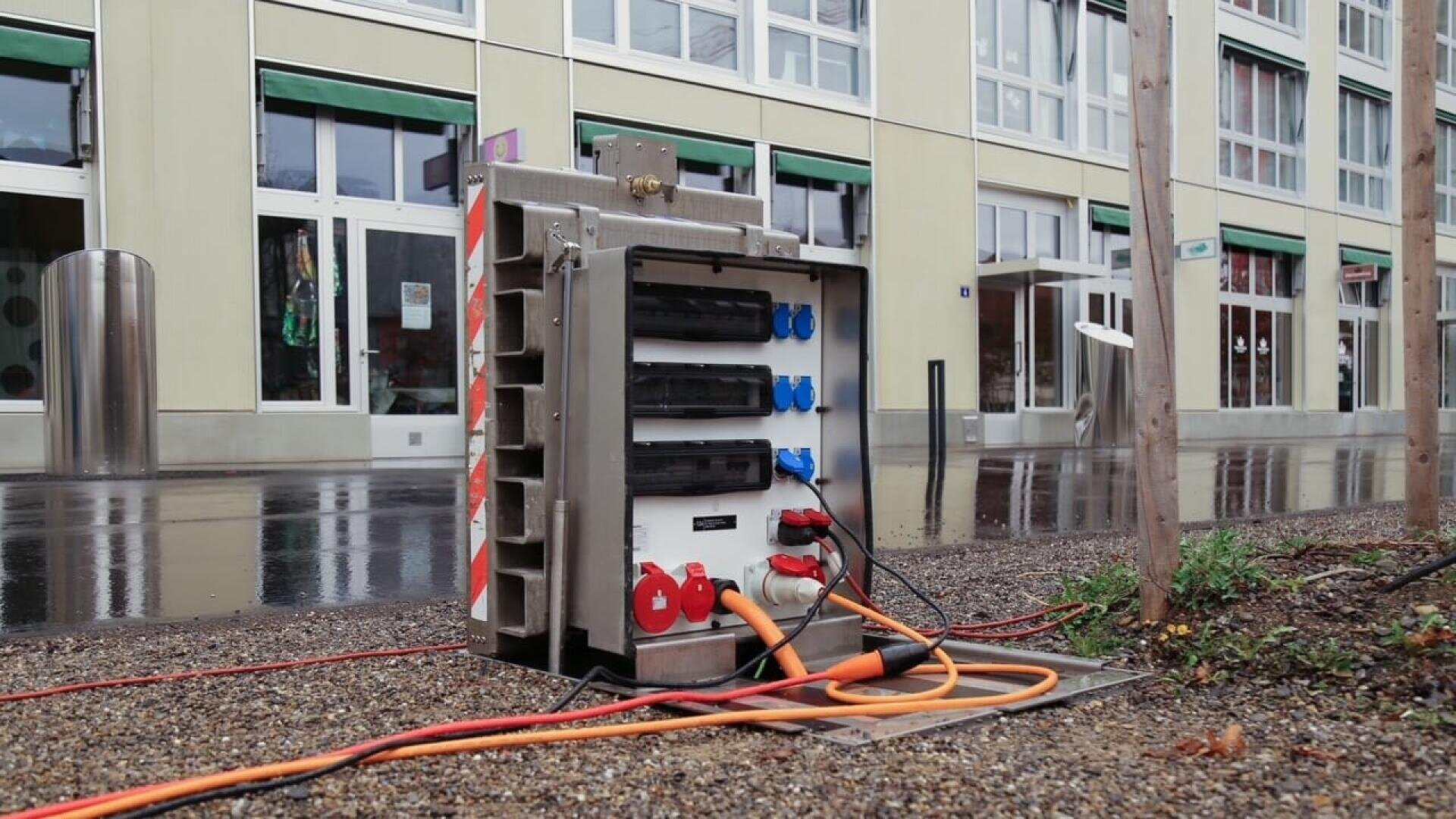 A portable electrical distribution box with multiple outlets and colored cables is set up outdoors on wet ground near a modern building with large windows and green awnings on a rainy day.