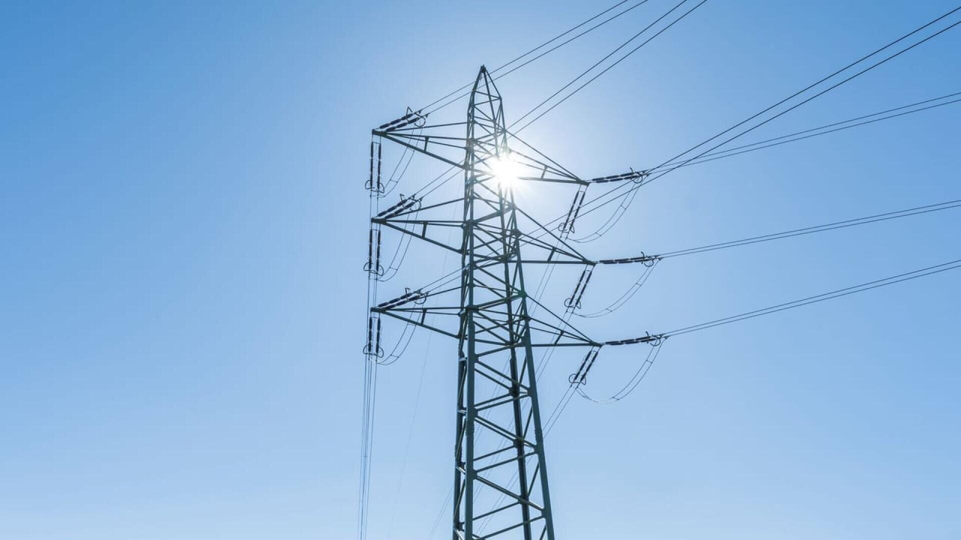 A tall electrical transmission tower with multiple power lines stretching outward, photographed from below against a clear blue sky with the sun shining behind the structure.
