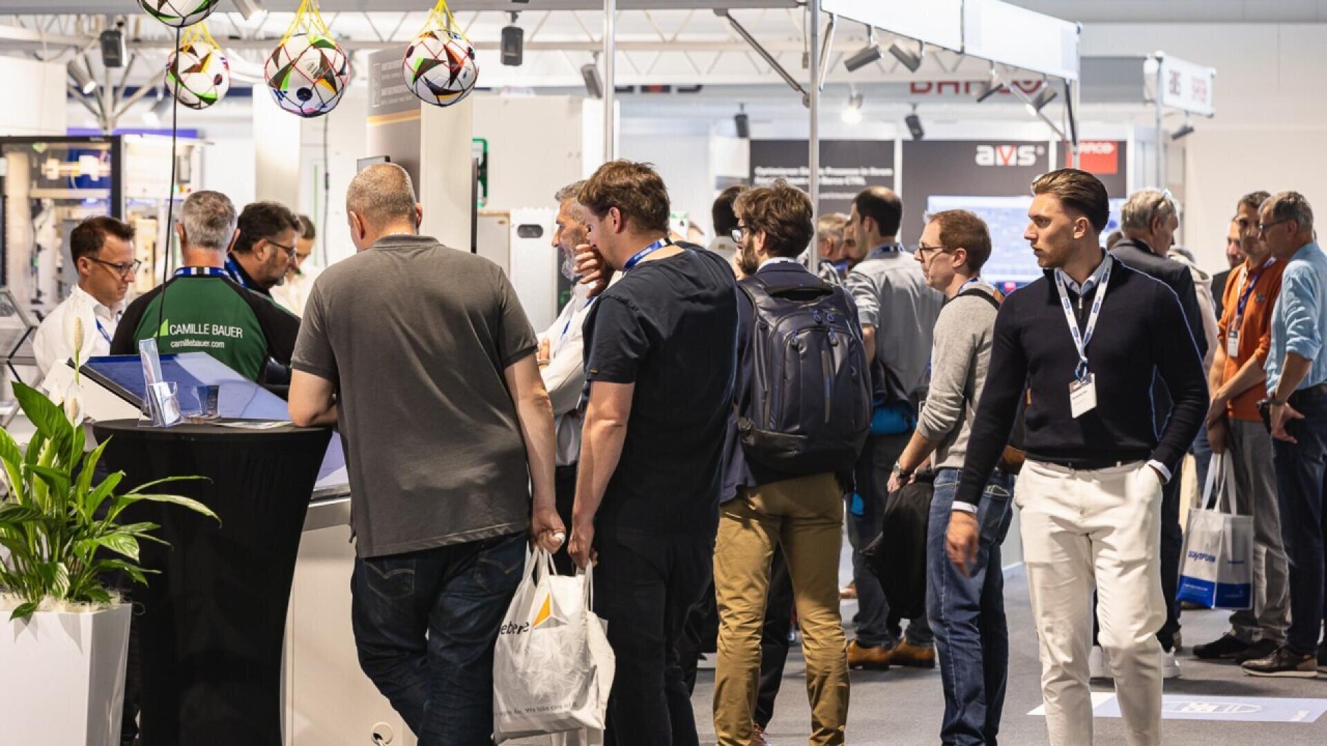 A group of people gather and interact at an indoor trade show or exhibition, some wearing lanyards and carrying bags. Booths and displays are visible in the background.