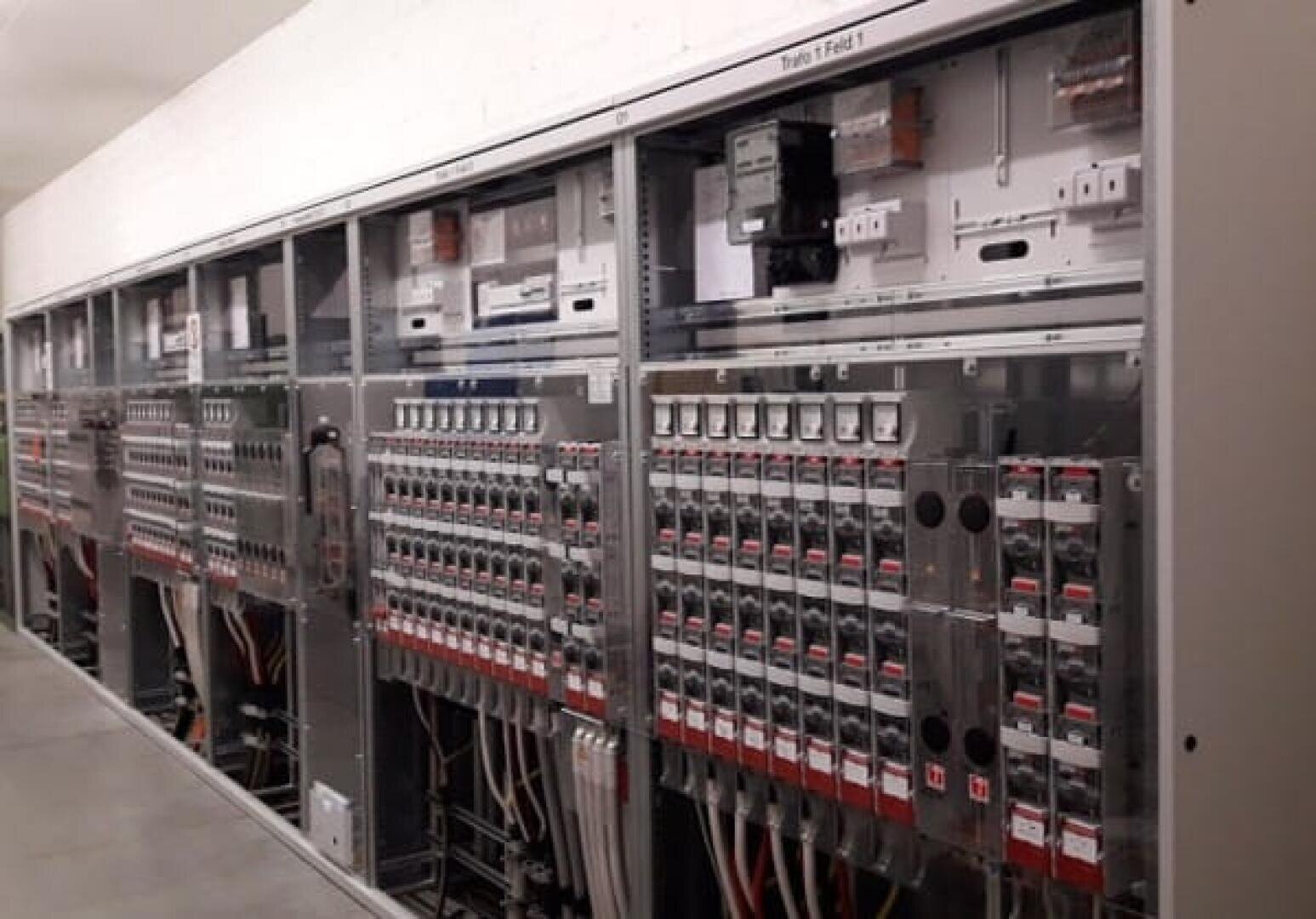 Eine Reihe elektrischer Schalttafeln in einem Versorgungsraum mit mehreren Schaltern, Leistungsschaltern, Kabeln und beschrifteten Bauteilen, die alle hinter transparenten Glastüren liegen.