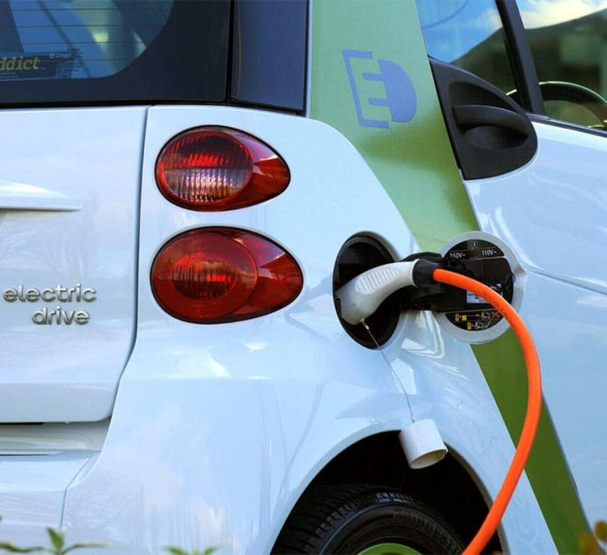 Ein weißes Elektroauto ist an eine Ladestation angeschlossen, wobei ein orangefarbenes Ladekabel mit dem Anschluss an der Rückseite des Fahrzeugs verbunden ist. Der Schriftzug "electric drive" ist auf dem Auto zu sehen.