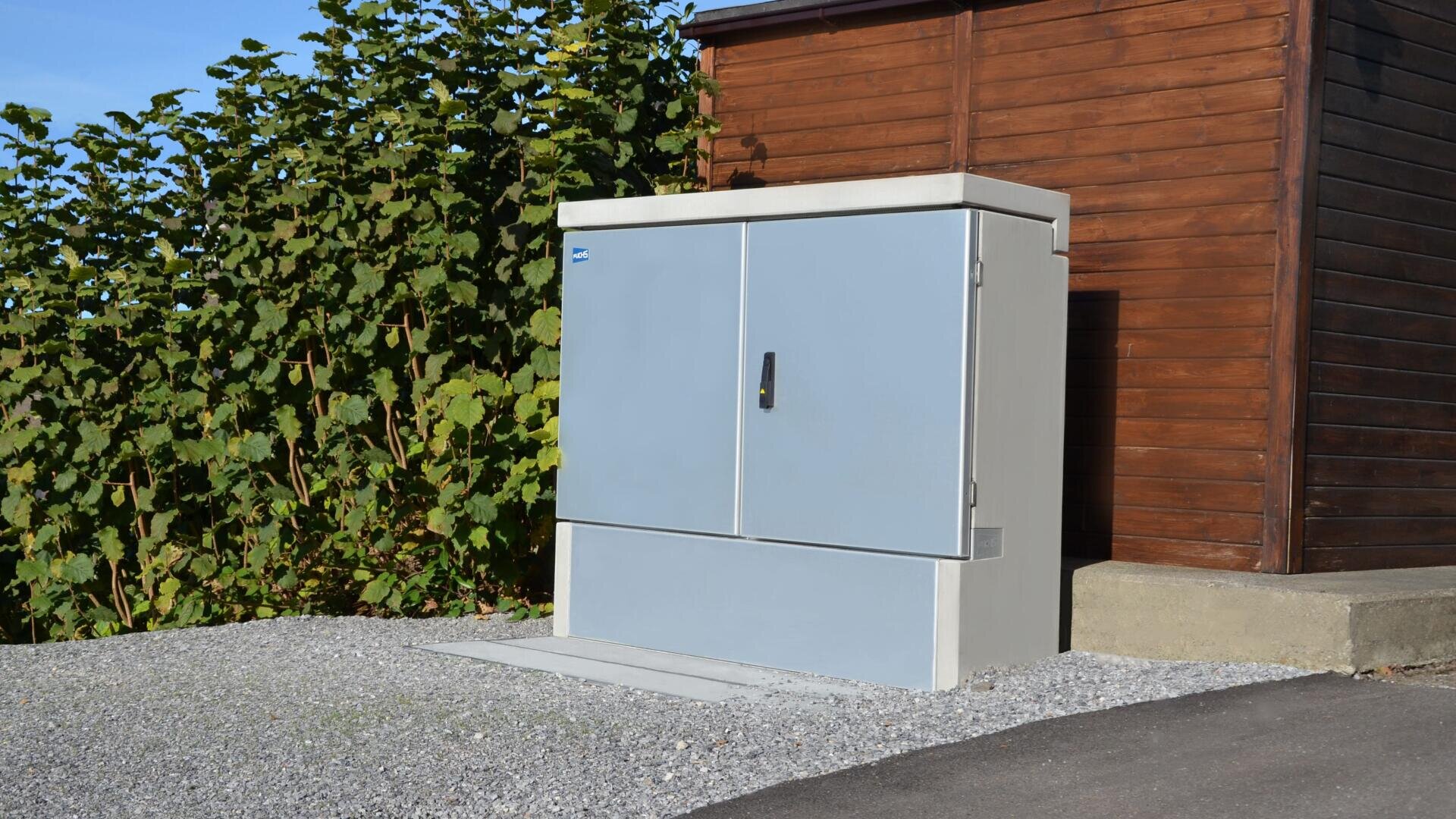 Ein großer grauer Stromkasten mit Flügeltüren steht auf einem Kiesplatz neben einem Holzgebäude und grünen Büschen, auf die das Sonnenlicht Schatten wirft.