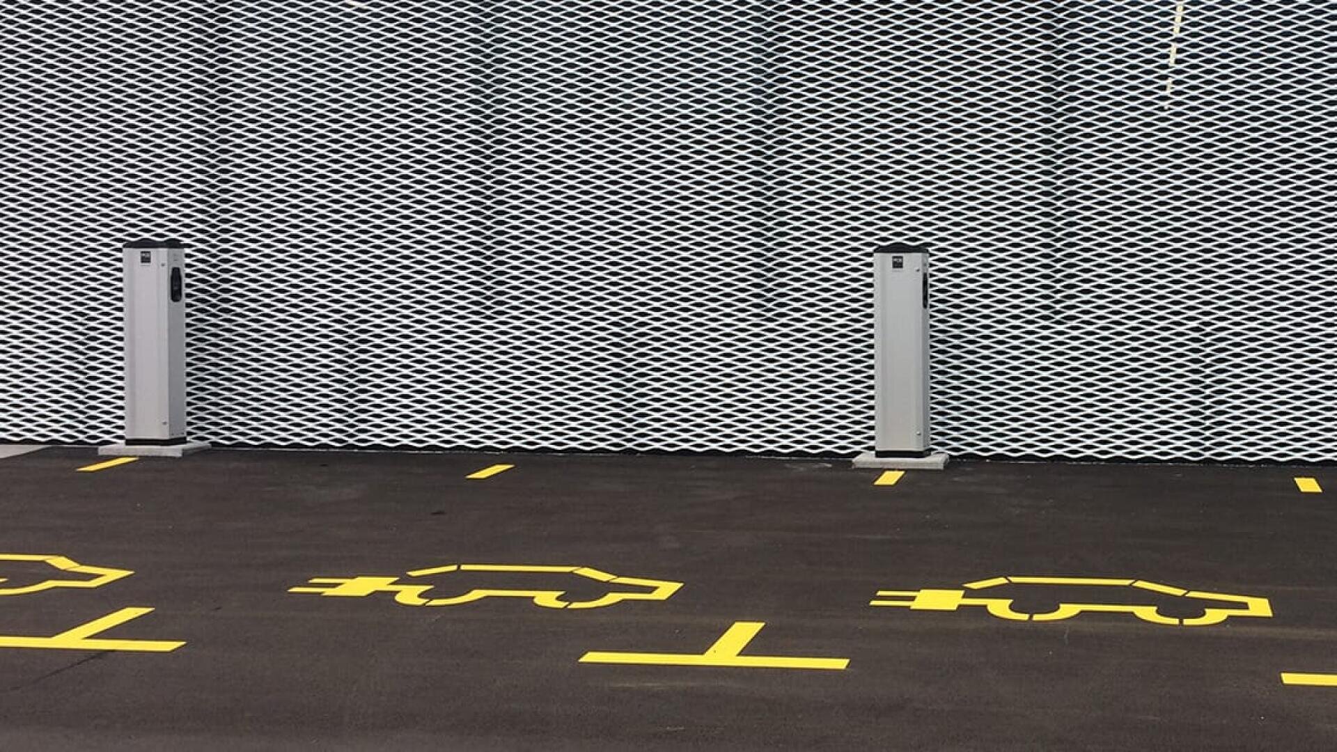 Drei Parkplätze mit gelben Elektroauto-Symbolen und zwei Ladestationen vor einem dunklen Bürgersteig und einem modernen, gemusterten weißen Metallzaun im Hintergrund.