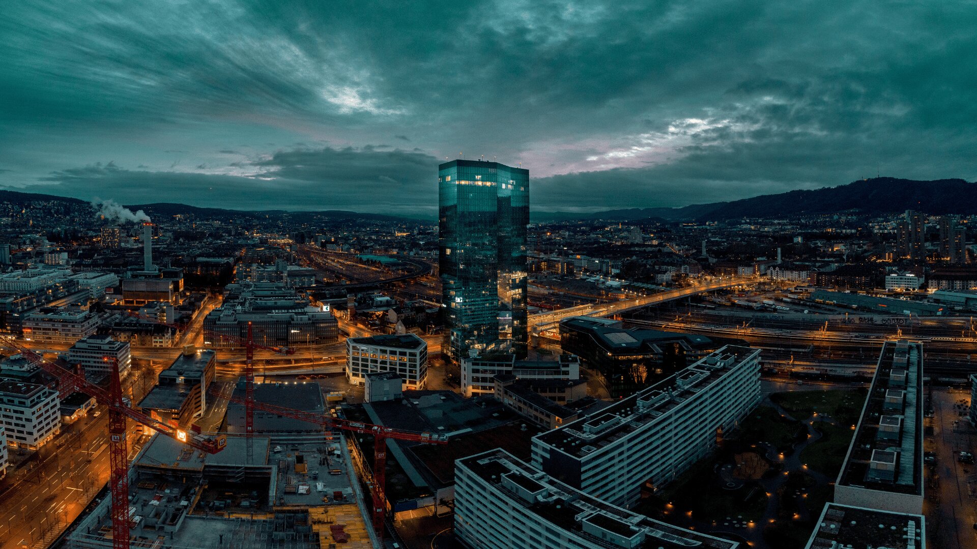 Aerial view of a city at dusk with a tall, glass skyscraper illuminated at the center, surrounded by modern buildings, roads, and railways, with mountains and a cloudy sky in the background.