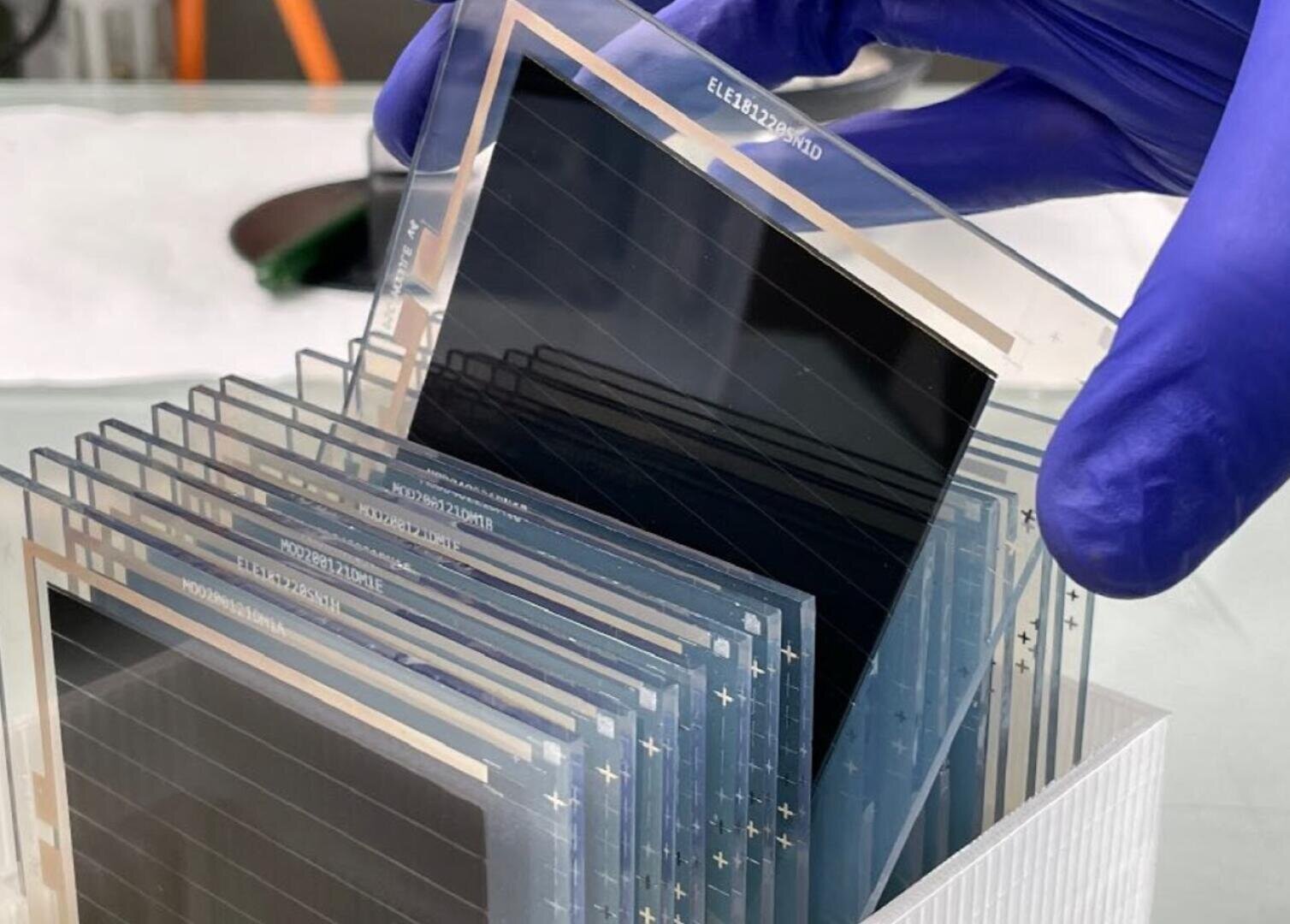 Eine behandschuhte Hand hält eine dünne, transparente Solarzelle oder elektronische Folie über einen Stapel ähnlicher rechteckiger Folien, die in einem Kunststoffhalter auf einem Labortisch angeordnet sind.