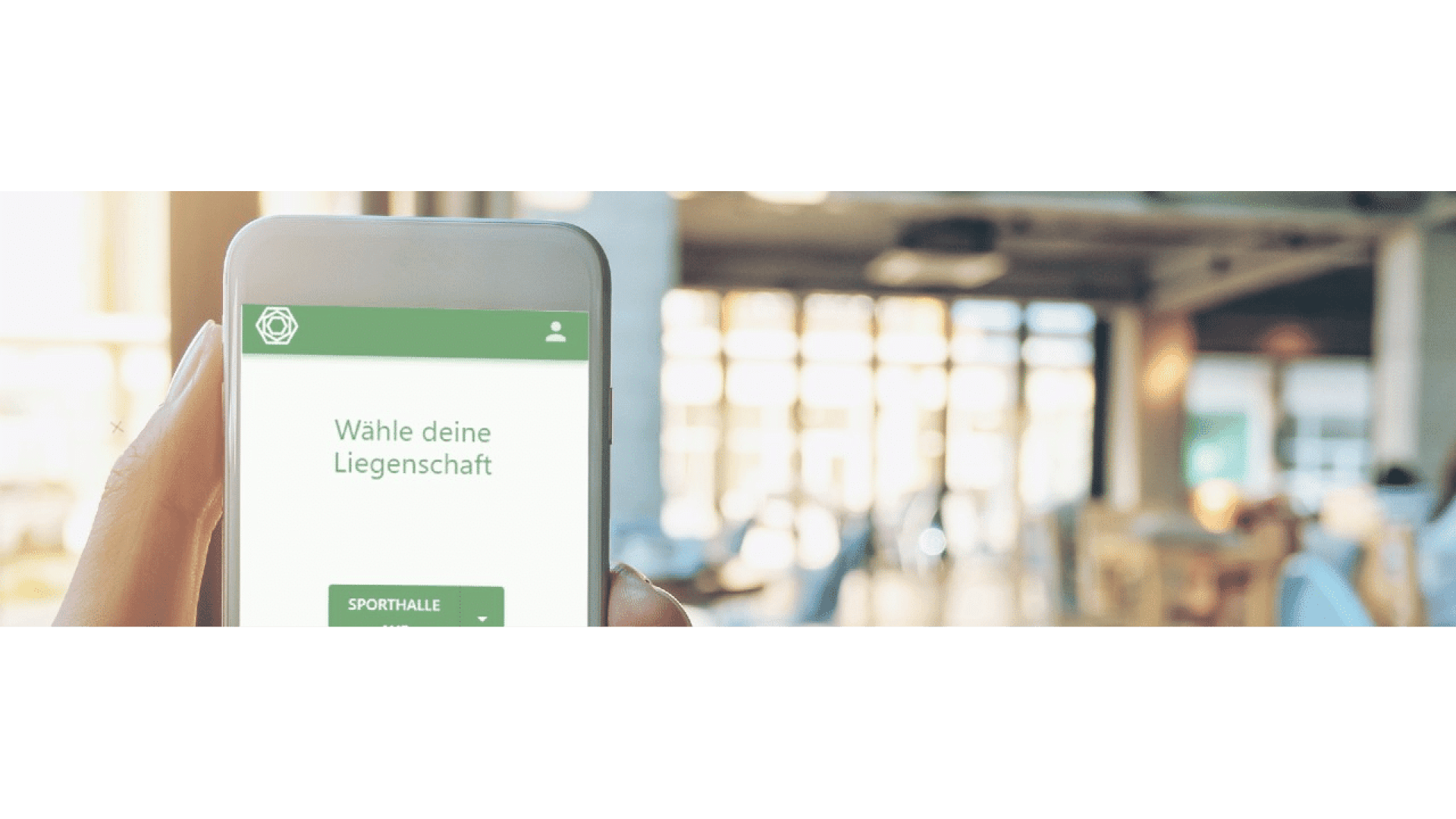 Eine Hand hält ein Smartphone, auf dem eine deutsche App mit der Aufschrift "Wähle deine Liegenschaft" angezeigt wird, während im Hintergrund ein unscharfes, sonnenbeschienenes Café oder Büro zu sehen ist.