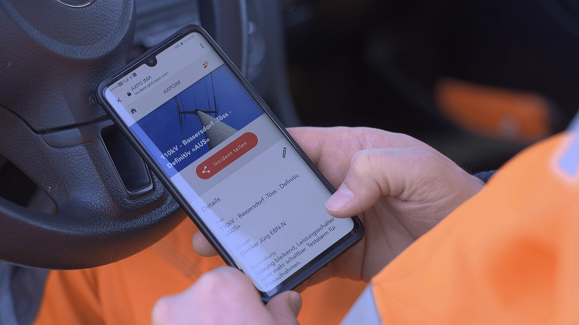 Eine Person, die eine orangefarbene Jacke trägt, benutzt ein Smartphone in einem Fahrzeug und hält das Telefon in beiden Händen in der Nähe des Lenkrads. Auf dem Bildschirm des Telefons wird eine Anwendung mit Text und einer roten Taste angezeigt.